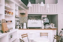 A cozy cafe interior featuring a countertop with a large espresso machine labeled 'Elektra'. The counter is adorned with a vase of flowers, various cups, and jars. Shelves on the left display an array of jams, honey, and tea, while a blender and other kitchen equipment are visible. The cafe is decorated in a soft, white color palette with wooden chairs and minimalistic decor.
