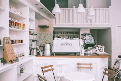 A cozy cafe interior featuring a countertop with a large espresso machine labeled 'Elektra'. The counter is adorned with a vase of flowers, various cups, and jars. Shelves on the left display an array of jams, honey, and tea, while a blender and other kitchen equipment are visible. The cafe is decorated in a soft, white color palette with wooden chairs and minimalistic decor.