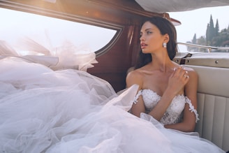 A woman in an elegant wedding dress with intricate lace details sits inside a wooden boat. Her long dark hair is loose, and she wears diamond earrings, creating a glamorous look. Soft morning light filters in, highlighting the fabric of her dress. The interior has a luxurious feel with cream-colored seats, and a serene outdoor scene with water and trees is visible through the open window.