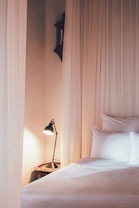A cozy bedroom setting features a neatly made bed with white pillows and sheets, softly illuminated by a black lamp on a bedside table. Sheer, light-colored curtains create a serene and inviting atmosphere.