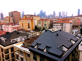 A densely packed urban neighborhood with various mid-rise residential buildings featuring a mix of flat and sloped roofs. The foreground includes a large building with a dark, geometric roof. Satellite dishes are visible on the rooftops. In the background, modern skyscrapers rise into a cloudy sky, indicating a city skyline.