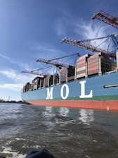 A large cargo ship is seen loaded with numerous shipping containers stacked neatly on its deck. The letters 'M O L' are prominently displayed on the side of the ship. Several cranes tower over the ship, indicating an active port environment. The water near the vessel is calm and a clear blue sky stretches overhead.