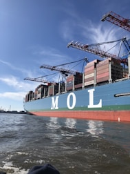 A large cargo ship is seen loaded with numerous shipping containers stacked neatly on its deck. The letters 'M O L' are prominently displayed on the side of the ship. Several cranes tower over the ship, indicating an active port environment. The water near the vessel is calm and a clear blue sky stretches overhead.