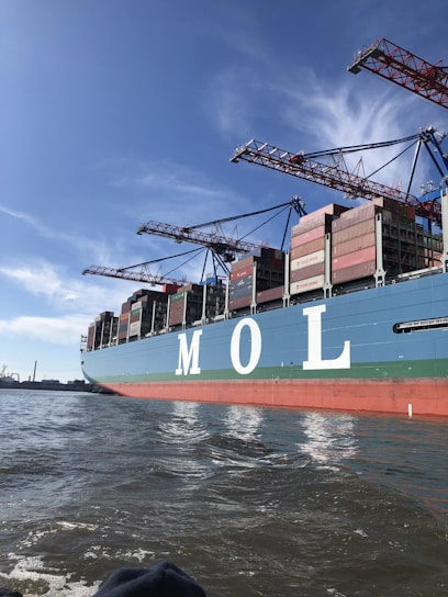 A large cargo ship is seen loaded with numerous shipping containers stacked neatly on its deck. The letters 'M O L' are prominently displayed on the side of the ship. Several cranes tower over the ship, indicating an active port environment. The water near the vessel is calm and a clear blue sky stretches overhead.