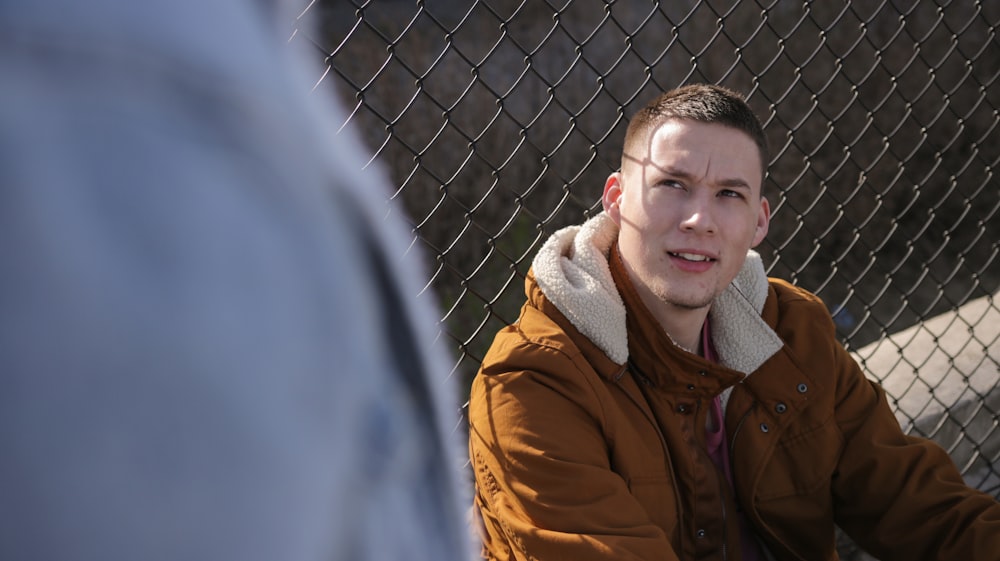 man wearing brown jacket sitting near gray link fence