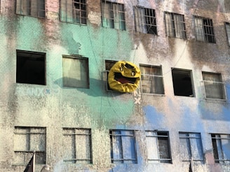 A building facade with a faded and weathered paint job. One window is creatively adorned with a large, crumpled sculpture resembling a yellow face with a wide-open mouth. The windows are partially open or covered with drapes.