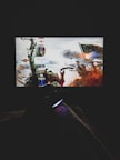 A dimly lit room with a television screen displaying a dynamic, action-packed video game interface featuring a historical battle scene. The foreground shows the silhouette of a gaming console controller held by a person.