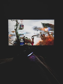A dimly lit room with a television screen displaying a dynamic, action-packed video game interface featuring a historical battle scene. The foreground shows the silhouette of a gaming console controller held by a person.