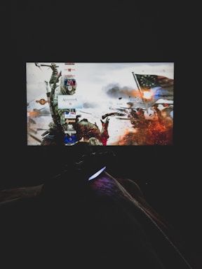 A dimly lit room with a television screen displaying a dynamic, action-packed video game interface featuring a historical battle scene. The foreground shows the silhouette of a gaming console controller held by a person.