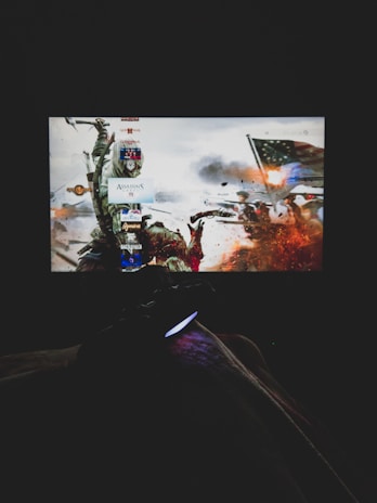 A dimly lit room with a television screen displaying a dynamic, action-packed video game interface featuring a historical battle scene. The foreground shows the silhouette of a gaming console controller held by a person.