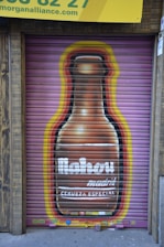 A painted mural on a metal shutter depicts a large beer bottle with the word 'Mahou' prominently displayed. The background features a gradient of colors, including yellow and red, surrounding the bottle. Above the mural is a yellow sign with partial text visible at the top.