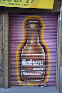 A painted mural on a metal shutter depicts a large beer bottle with the word 'Mahou' prominently displayed. The background features a gradient of colors, including yellow and red, surrounding the bottle. Above the mural is a yellow sign with partial text visible at the top.