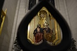 A religious sculpture is seen through a large, stylized heart-shaped frame. The sculpture features two seated figures adorned in flowing robes in an ornate setting with rich detailing and vibrant colors. The backdrop consists of an intricate patterned design in gold and green tones, complemented by surrounding dark foliage.
