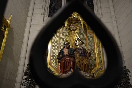 A religious sculpture is seen through a large, stylized heart-shaped frame. The sculpture features two seated figures adorned in flowing robes in an ornate setting with rich detailing and vibrant colors. The backdrop consists of an intricate patterned design in gold and green tones, complemented by surrounding dark foliage.