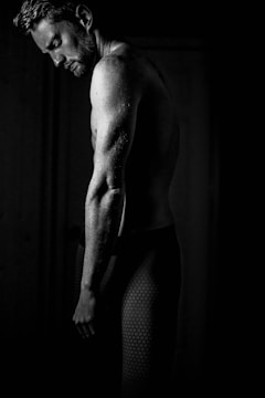 A black and white portrait of a man captured in profile, his upper body slightly turned and head bent forward, casting gentle shadows that accentuate his muscular frame. The textured pants he wears have a hexagonal pattern, adding a subtle contrast to the soft lighting.