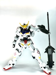 A white and red humanoid robot figure with yellow and blue accents, holding a large black weapon. It has a sleek and futuristic design with prominent armor plating and intricate detailing, standing upright against a plain background.