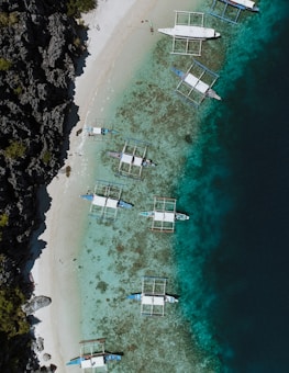 An aerial view of a beach with several traditional boats anchored near the shore. The clear turquoise waters reveal coral formations below, contrasting with the deeper blue of the ocean further out. A rocky cliff with sparse greenery lines the left side of the image, while a narrow sandy beach runs parallel to the water.