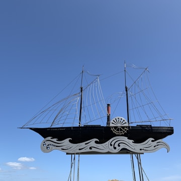 A stylized sculpture of a ship is suspended against a clear blue sky. The ship features ironwork detailing with intricate waves at the bottom and wireframe sails above. The overall design is minimalist and artistic, emphasizing form over detail.