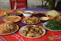 A variety of dishes are displayed on a colorful tablecloth, including stuffed rolls, fried snacks, leafy green vegetables, a bowl of dried fruits, and other mixed plates. Some dishes are served in ceramic bowls and surrounded by a natural outdoor setting.