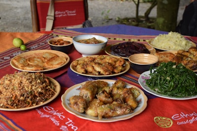A variety of dishes are displayed on a colorful tablecloth, including stuffed rolls, fried snacks, leafy green vegetables, a bowl of dried fruits, and other mixed plates. Some dishes are served in ceramic bowls and surrounded by a natural outdoor setting.