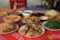 A variety of dishes are displayed on a colorful tablecloth, including stuffed rolls, fried snacks, leafy green vegetables, a bowl of dried fruits, and other mixed plates. Some dishes are served in ceramic bowls and surrounded by a natural outdoor setting.