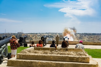A group of people are observing a historical reenactment with cannons situated on a grassy area overlooking a cityscape. Smoke is billowing from one of the cannons as it has just been fired. The sky is clear with some clouds, and stone steps lead up to the area where the cannons are displayed.