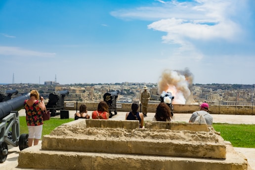 A group of people are observing a historical reenactment with cannons situated on a grassy area overlooking a cityscape. Smoke is billowing from one of the cannons as it has just been fired. The sky is clear with some clouds, and stone steps lead up to the area where the cannons are displayed.