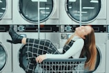 A woman is comfortably sitting and laughing inside a laundry cart, positioned in front of rows of industrial washing machines in a laundromat. She is wearing a black and white checkered outfit with black heeled boots.
