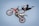 A person is performing an acrobatic stunt in the air on a dirt bike. The rider is wearing protective gear including a helmet and boots, with one leg extended behind and both hands gripping the handlebars. The background is clear and blue, emphasizing the height and skill involved in the trick.