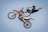 A person is performing an acrobatic stunt in the air on a dirt bike. The rider is wearing protective gear including a helmet and boots, with one leg extended behind and both hands gripping the handlebars. The background is clear and blue, emphasizing the height and skill involved in the trick.