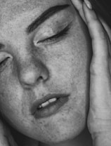 A close-up black and white photograph of a person's face with closed eyes. The person has freckles and is gently holding their face with both hands, conveying a sense of calm or introspection.
