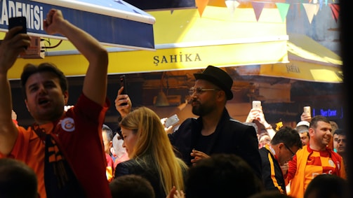 A lively crowd gathers in an outdoor setting with many people capturing the moment on their phones. The atmosphere is cheerful and energetic, with some individuals wearing sports jerseys, likely celebrating or supporting an event.