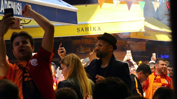 A lively crowd gathers in an outdoor setting with many people capturing the moment on their phones. The atmosphere is cheerful and energetic, with some individuals wearing sports jerseys, likely celebrating or supporting an event.