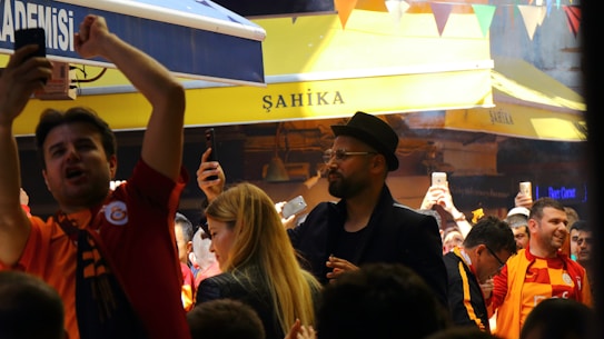 A lively crowd gathers in an outdoor setting with many people capturing the moment on their phones. The atmosphere is cheerful and energetic, with some individuals wearing sports jerseys, likely celebrating or supporting an event.