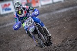 A motocross rider is captured racing through a muddy track with determination. The rider is dressed in a colorful suit with logos, wearing a helmet with a visor. The motorcycle is splattered with mud, and the scene is dynamic with dirt flying around.