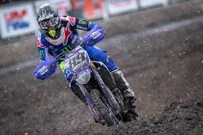 A motocross rider is captured racing through a muddy track with determination. The rider is dressed in a colorful suit with logos, wearing a helmet with a visor. The motorcycle is splattered with mud, and the scene is dynamic with dirt flying around.