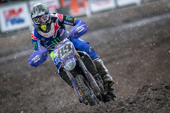 A motocross rider is captured racing through a muddy track with determination. The rider is dressed in a colorful suit with logos, wearing a helmet with a visor. The motorcycle is splattered with mud, and the scene is dynamic with dirt flying around.