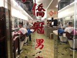 A barbershop scene features a barber wearing a mask, working on a customer's hair. Reflections in mirrors create a sense of depth, and Chinese characters are prominently displayed on the glass door. Hair clippings are visible on the tiled floor, and the interior gives a retro vibe.