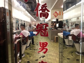 A barbershop scene features a barber wearing a mask, working on a customer's hair. Reflections in mirrors create a sense of depth, and Chinese characters are prominently displayed on the glass door. Hair clippings are visible on the tiled floor, and the interior gives a retro vibe.