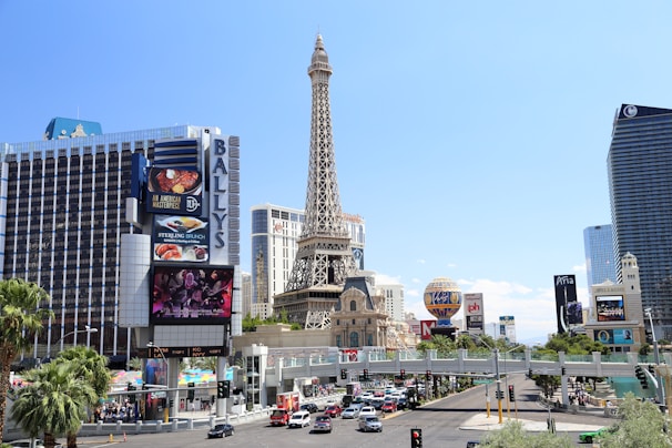 A lively urban scene featuring large hotels, including a tall structure resembling the Eiffel Tower, surrounded by palm trees and numerous advertisement billboards. Below, cars and pedestrians move along a busy street.
