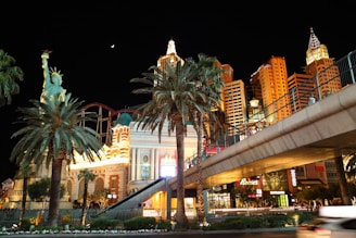 A nighttime scene featuring a replica of the Statue of Liberty surrounded by palm trees at the New York-New York Hotel & Casino. The backdrop includes a collection of illuminated buildings designed to resemble the New York City skyline. A pedestrian bridge crosses above a road, and colorful lights and advertisements are visible.