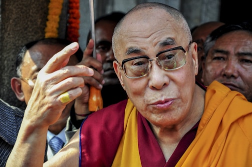 An elderly person in glasses, wearing a maroon and yellow robe, is surrounded by others. The focus appears to be on an expressive gesture made with the hands. In the background, people are closely positioned, some partially obscured.