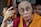 An elderly person in glasses, wearing a maroon and yellow robe, is surrounded by others. The focus appears to be on an expressive gesture made with the hands. In the background, people are closely positioned, some partially obscured.