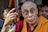 An elderly person in glasses, wearing a maroon and yellow robe, is surrounded by others. The focus appears to be on an expressive gesture made with the hands. In the background, people are closely positioned, some partially obscured.