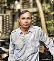 A man wearing a light blue uniform shirt with 'Mahadev Travels' embroidered on the pocket stands outdoors. He is leaning against a Hero Honda motorcycle with a blurred background of a street scene.