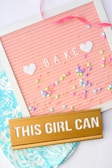A pink letter board with white letters spelling 'BAKE' adorned with two white heart shapes and colorful beads scattered around. A gold plaque stating 'THIS GIRL CAN' lies on top of a patterned blue fabric. A pink ribbon is loosely draped across the top corner.