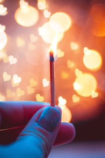 A hand holding a lit matchstick against a warm, softly focused background featuring bokeh lights shaped like hearts.