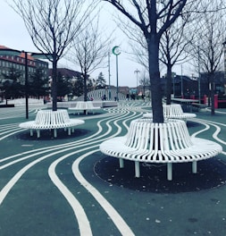 An urban park environment with unique design features, including wavy white lines on the ground that curve around circular benches constructed of white slats. These benches encircle several leafless trees. In the distance, there are buildings with red-tiled roofs and people walking around. The overall setting is modern with an artistic flair, creating a visually interesting landscape.