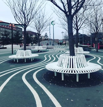 An urban park environment with unique design features, including wavy white lines on the ground that curve around circular benches constructed of white slats. These benches encircle several leafless trees. In the distance, there are buildings with red-tiled roofs and people walking around. The overall setting is modern with an artistic flair, creating a visually interesting landscape.