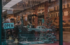A barber shop with a festive Christmas tree inside, decorated with red ornaments. A barber is working on a customer's haircut. The shop's name and the word 'OPEN' are visible on the glass window. The interior showcases a warm, rustic decor with wooden walls and ambient lighting.